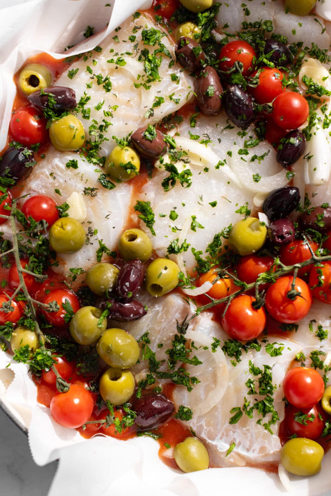up close braiser with uncooked mediterranean fish - thick chunks of cod in a herb simmered red sauce with olives and tomatoes.