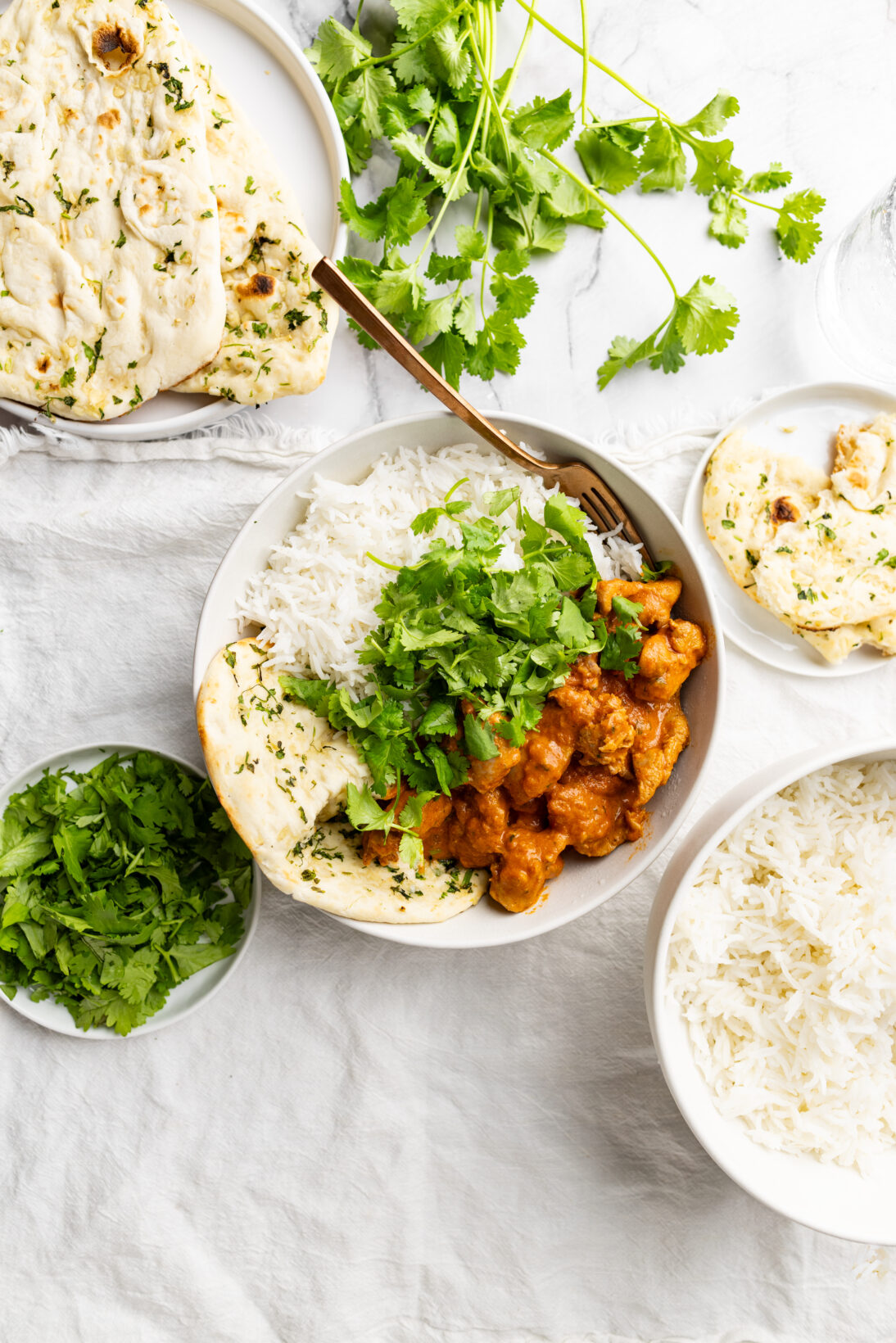bowl of butter chicken next to basmati rice and garlic naan, topped with roughly chopped cilantro