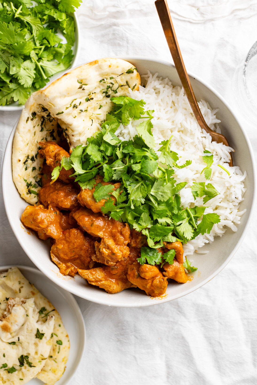 bowl of butter chicken next to basmati rice and garlic naan, topped with roughly chopped cilantro