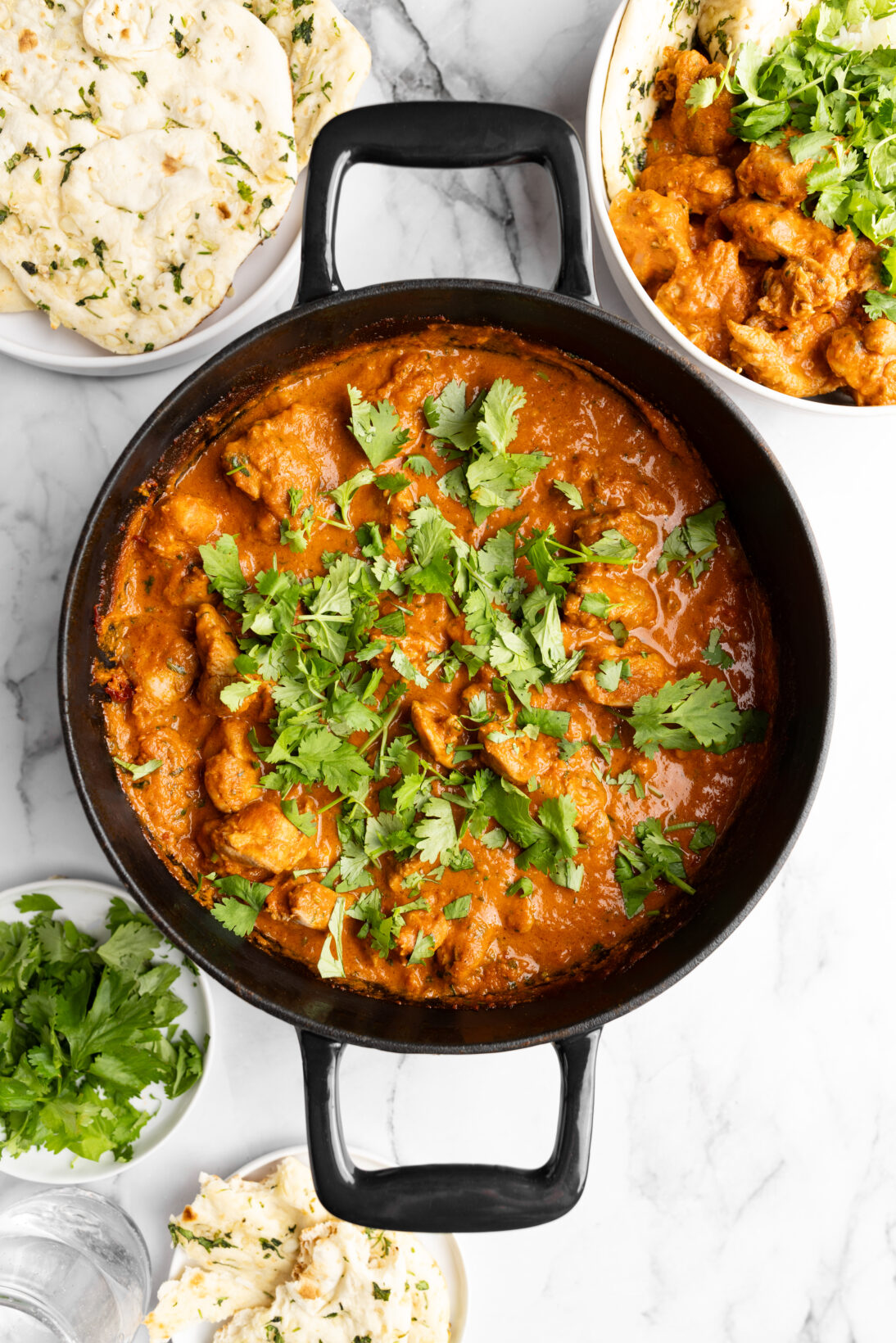 cast iron of butter chicken next to basmati rice and garlic naan, topped with roughly chopped cilantro