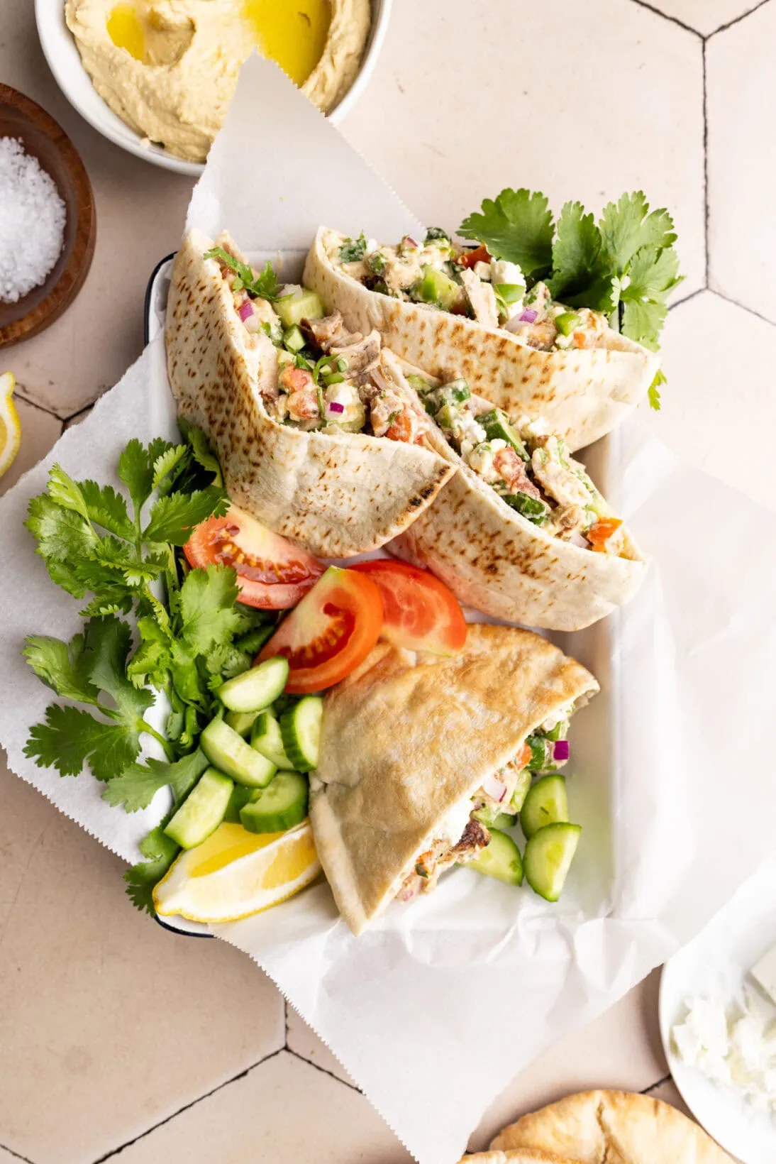 a tray of pita bread stuffed with hummus chicken salad. Also in the tray is bunches of cilantro, tomatoes, and lemon.
