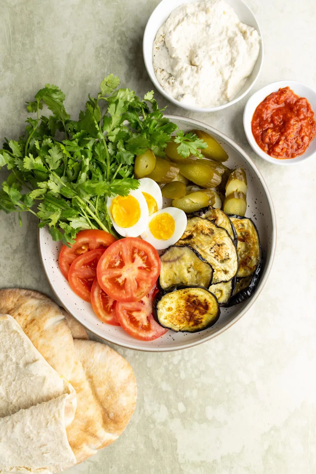 the ingredients for sabich sandwich ( roasted eggplant, hard boiled eggs, tomatoes,cilantro, cucumbers, baba ganoush, and harissa)