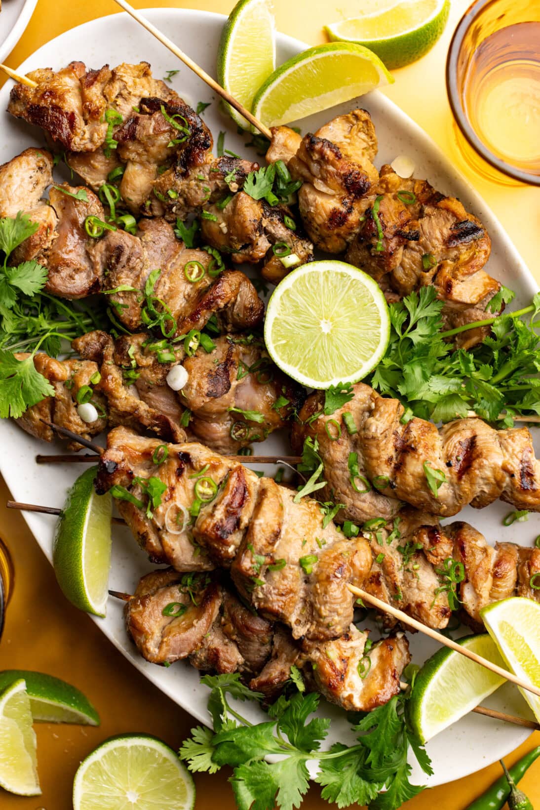 a serving plate of moo ping skewers topped with chilies, cilantro, green onions and limes.