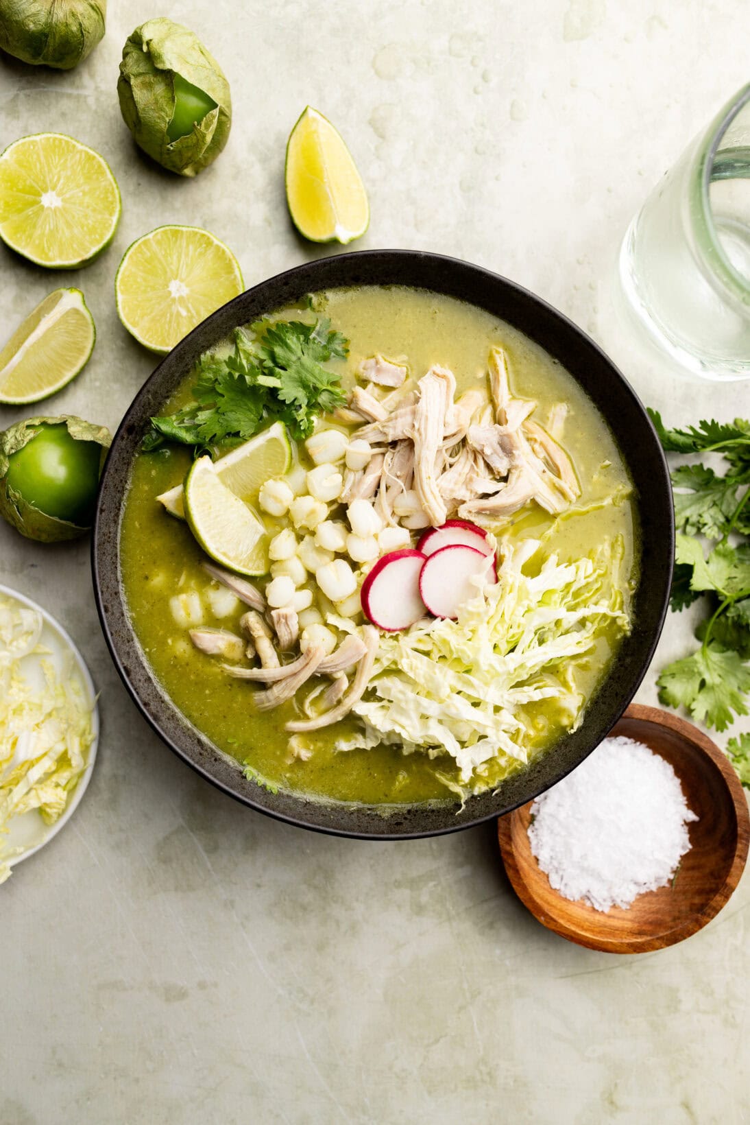 a bowl of pozole verde topped with extra cilantro, radish, cabbage, and wedges of lime