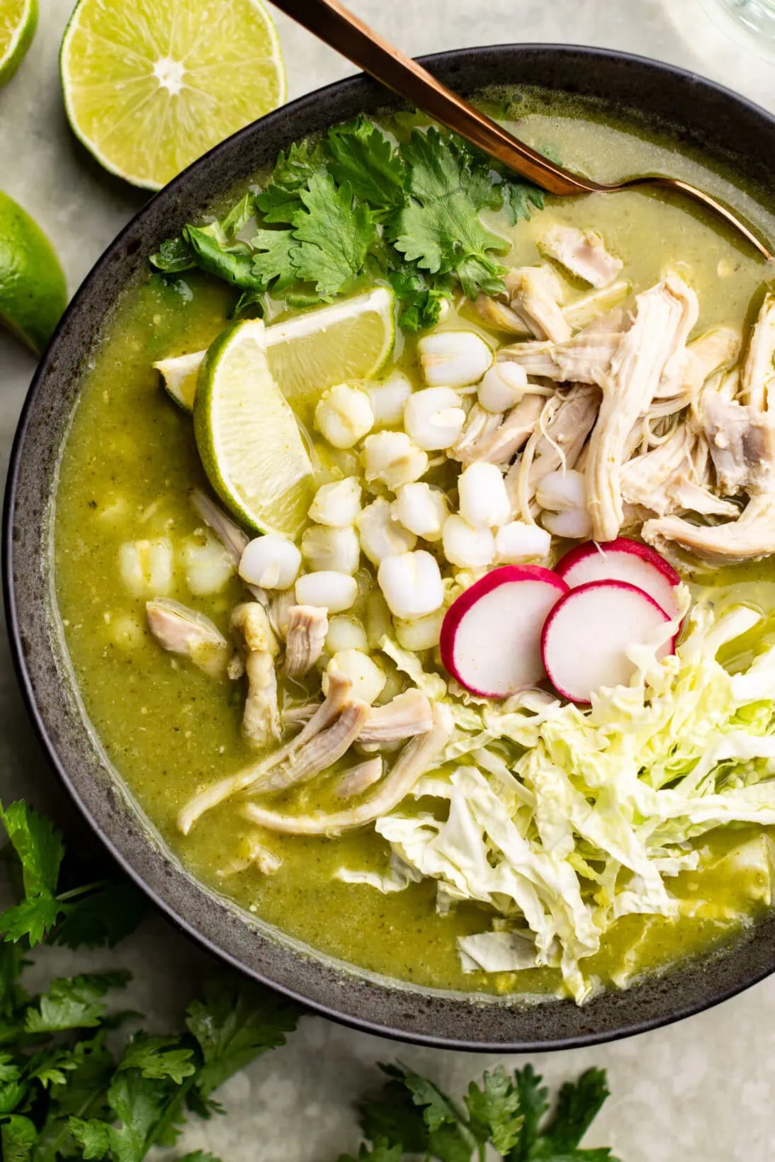 a bowl of pozole verde topped with extra cilantro, radish, cabbage, and wedges of lime