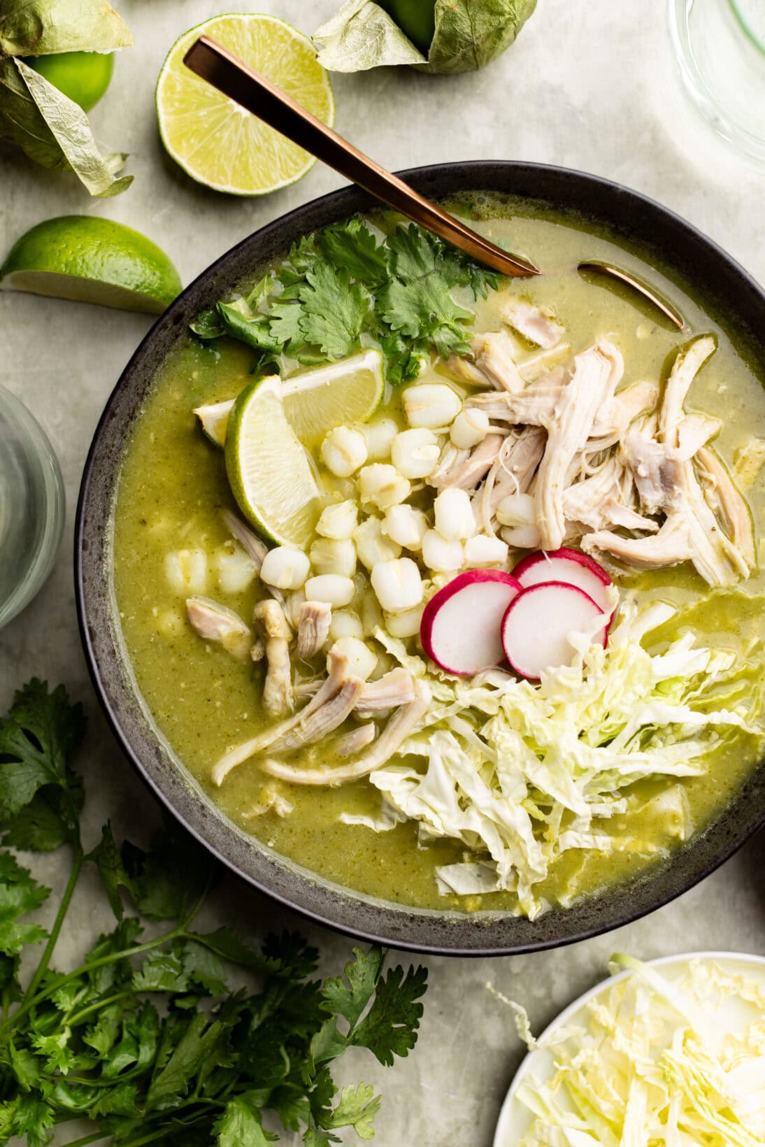 a bowl of pozole verde topped with extra cilantro, radish, cabbage, and wedges of lime