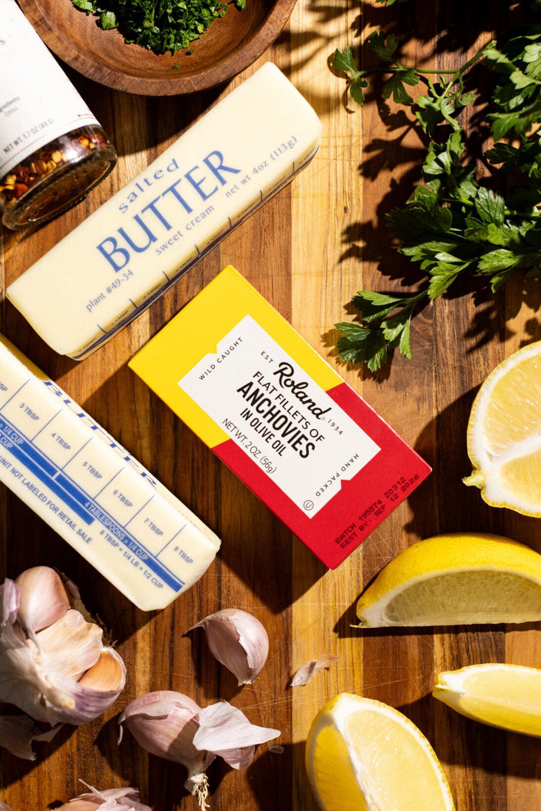 a box of anchovies surrounded by the other ingredients: parsley, chili flakes, lemons, and garlic.