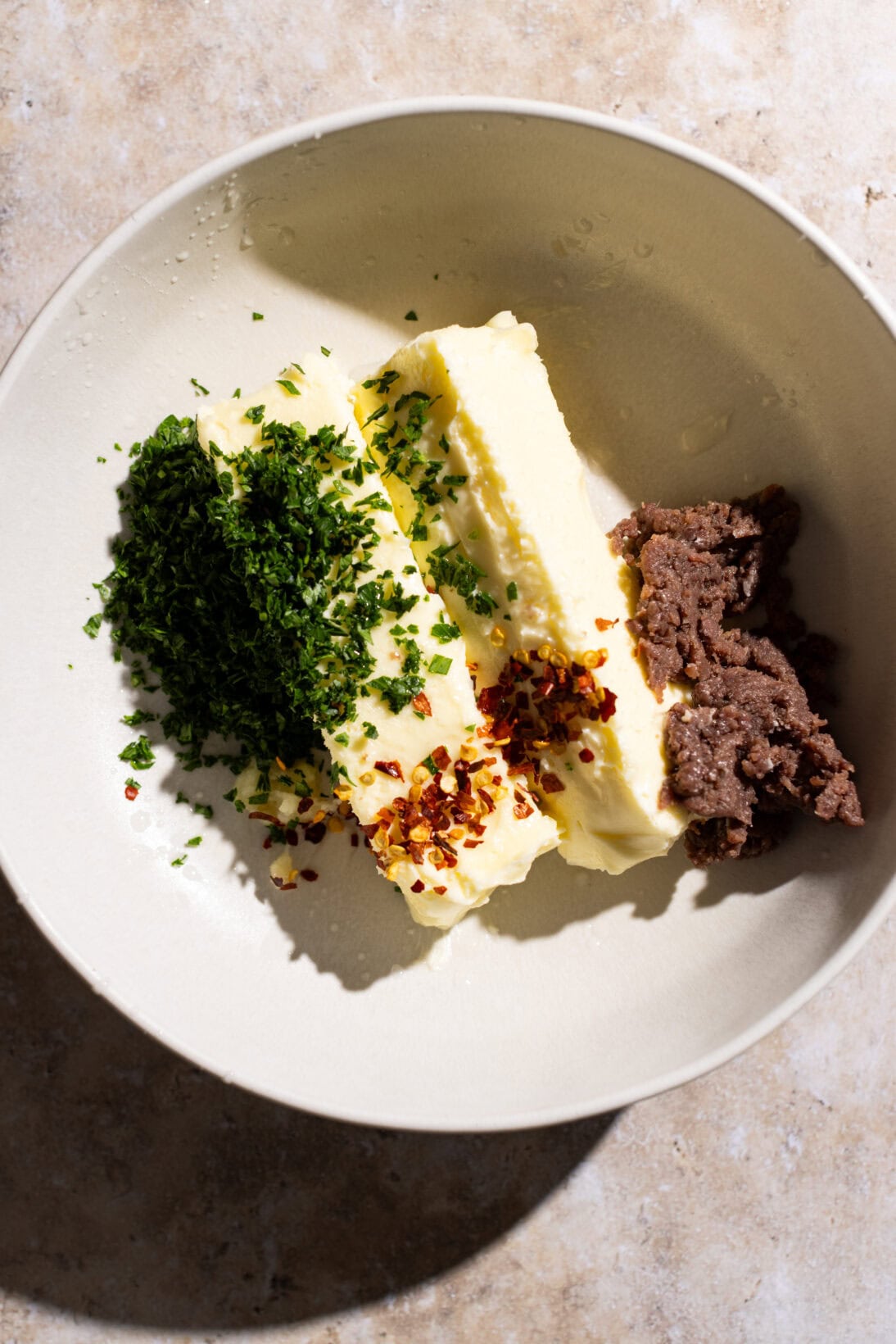 step by step: ingredients added to a bowl: anchovy paste, butter, chili flakes, parsley, and garlic.