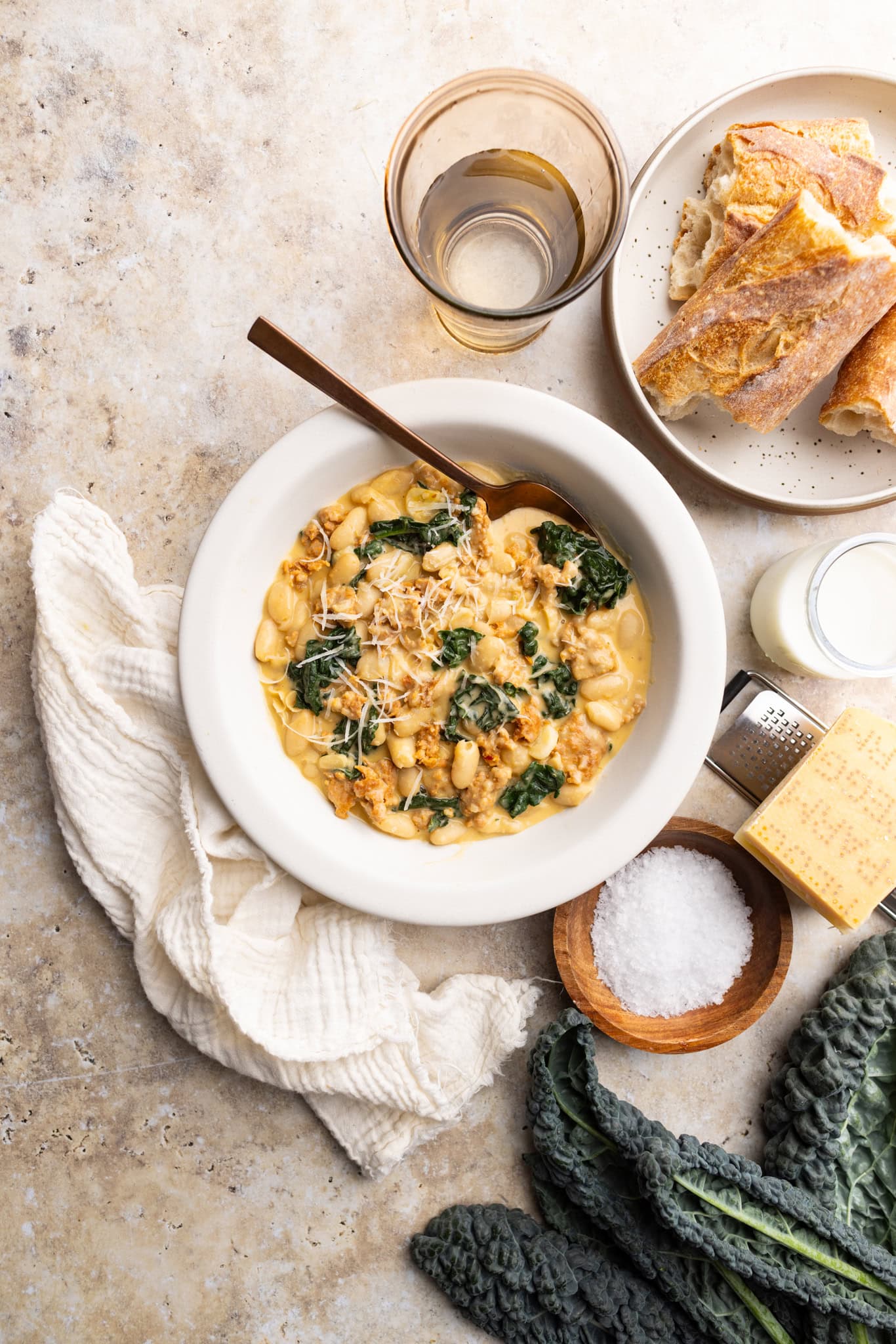 A bowl of Parmesan tuscan white beans with hot Italian sausage and kale. Topped with extra grated Parmesan. Surrounded by the ingredients and a bowl of crusty bread.