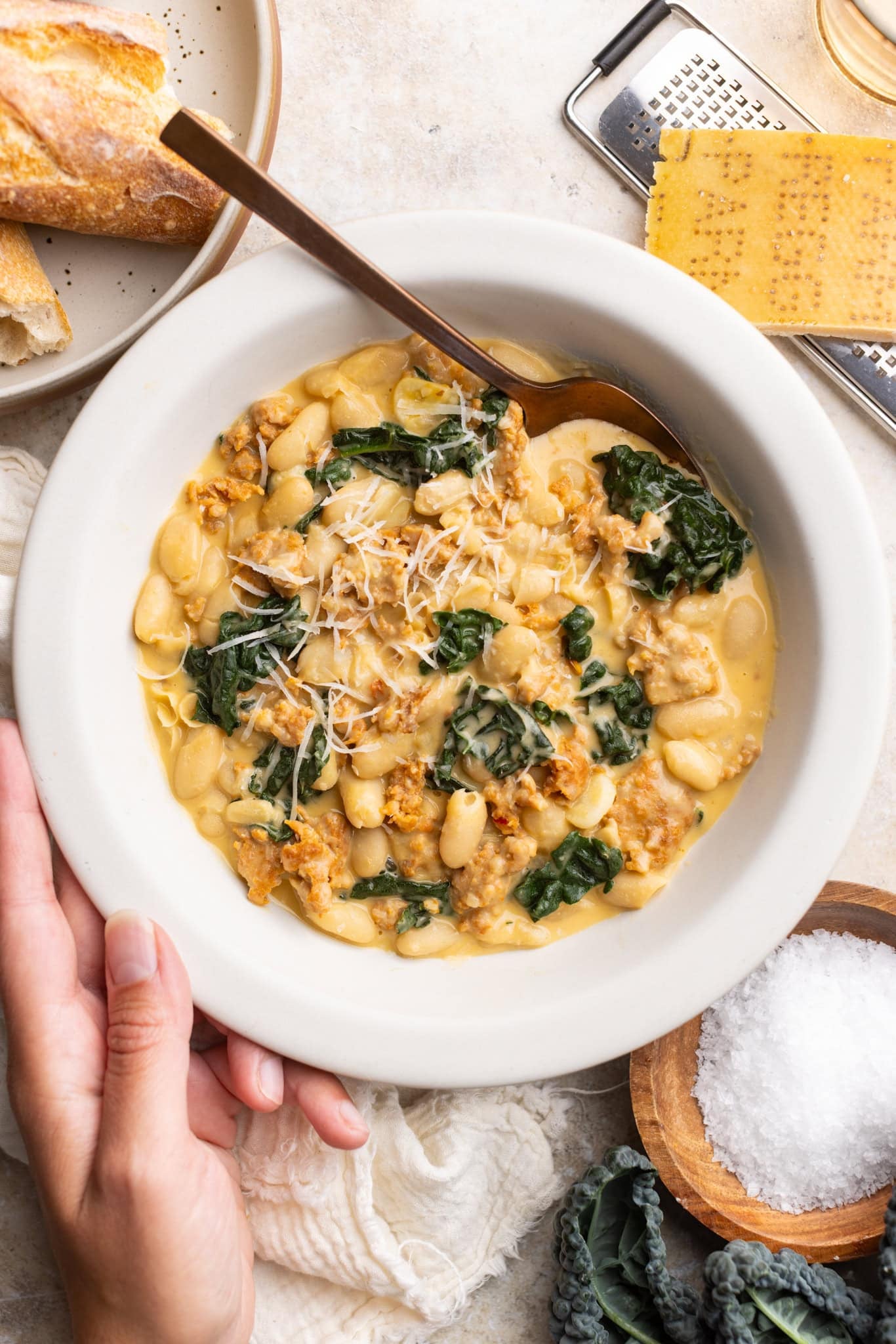 one hand holding a bowl of Parmesan tuscan white beans with hot Italian sausage and kale. Topped with extra grated Parmesan.